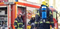 Three firefighters in full gear, including oxygen tanks, stand near a fire truck with its compartments open. The scene appears to be in an urban setting with buildings in the background. The firefighters, likely associates discussing a strategy or engaged in activity, convey a sense of readiness and teamwork.