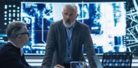 A group of five people are in a meeting room with a large digital screen showing data and maps in the background. A man with gray hair and beard stands and speaks to the seated individuals, who are attentively listening. The room has a futuristic, high-tech ambiance.